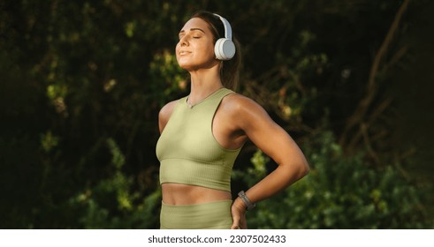 Woman listening to music and doing a breathing exercise outdoors. Woman meditating while standing against a nature background. Sports woman warming up for a morning workout. - Powered by Shutterstock