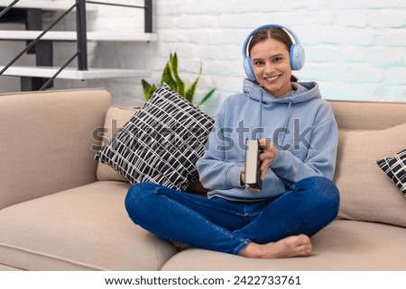 Woman listening music from headphones and reading book while sitting on sofa in living room.