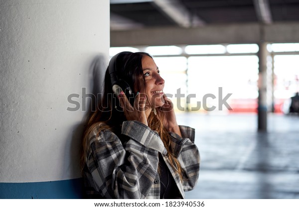 Woman listening to music with\
headphones. She is leaning against the column of a parking\
lot.