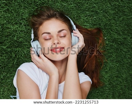 Woman listening to music with headphones in a white T-shirt on the green lawn grass in the park, vacation smile with teeth, looking into the camera