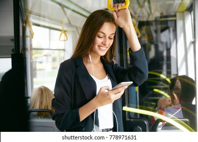 Woman Listening Music On Phone Riding In Bus. Portrait Of Stylish Smiling Girl Listening Music In Headphones, Using Smartphone While Riding In Public Transport. High Resolution.