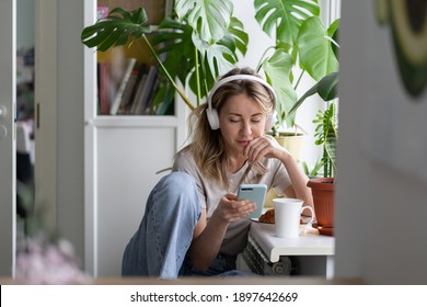 Woman listening to music, wear wireless white headphones, using mobile smart phone, chatting in social networks, sitting next to the window, houseplants on windowsill. Life at home. Time to relax. 