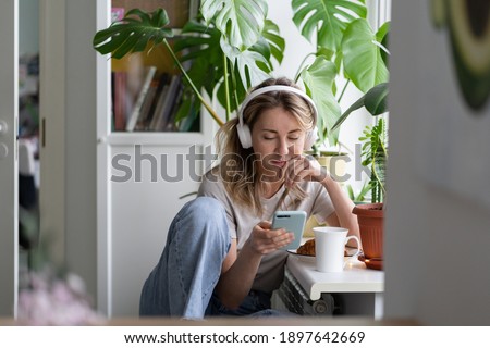 Woman listening to music, wear wireless white headphones, using mobile smart phone, chatting in social networks, sitting next to the window, houseplants on windowsill. Life at home. Time to relax. 
