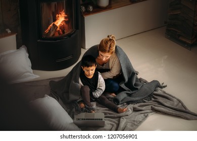 Woman and a little boy with blanket sitting on floor looking at  laptop. Mother and son watching cartoons on laptop sitting near fireplace at home. - Powered by Shutterstock