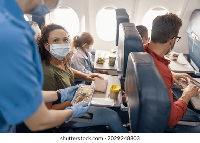 Woman With Little Daughter Wearing Protective Face Mask Looking At Female Flight Attendant Serving Lunch To Passengers On Board. Traveling By Airplane During Covid19