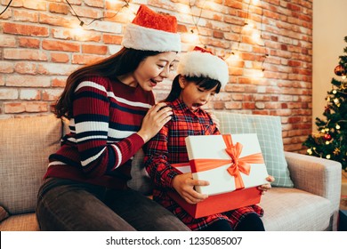 Woman And Little Girl Curiously Open Big Red Gift Box On Boxing Day. Family Wearing Santa Claus Hats Celebrating Xmas In Cozy Decorated Room At Home. Christmas Present Suprised Concept.