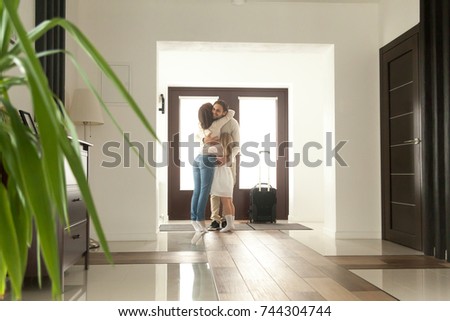Woman and little girl embracing man standing in hall, wife meeting husband just arrived came home with travel case returning after business trip, happy family hugging father, welcome back sweet home 