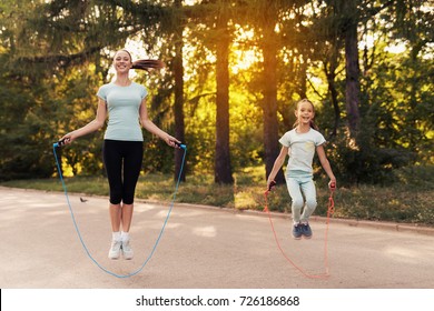 A woman and a little girl are jumping rope on the path in the park. They are having fun