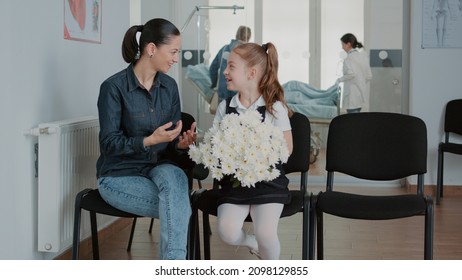 Woman And Little Girl Preparing To Visit Patient In Waiting Room, Having Flowers For Sick Person. Mother And Child Waiting To Enter Hospital Ward At Medical Facility. People In Waiting Area