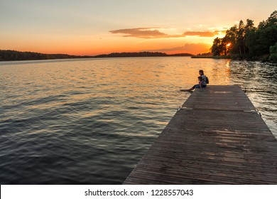 Eine Frau und ein kleines Mädchen sitzen auf dem Steg, und ihre nackten Füße schwingen im See. Die Vereinigung von Mutter und Tochter. Schöner Sonnenuntergang mit warmem, weichem Licht.