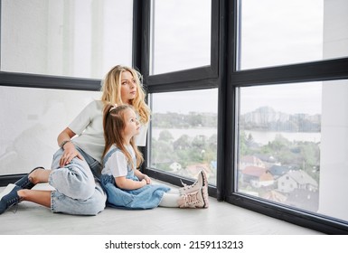 Woman And Little Girl Sitting On The Floor And Looking Out Window In Family New Home. Mother And Child Enjoying View From Panoramic Window In Newly Built Apartment.