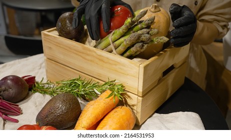 Woman Loads Wooden Box With Organic Vegetables. Female Hands In Black Rubber Gloves Put Fresh Vegetables Into Small Wooden Box Closeup