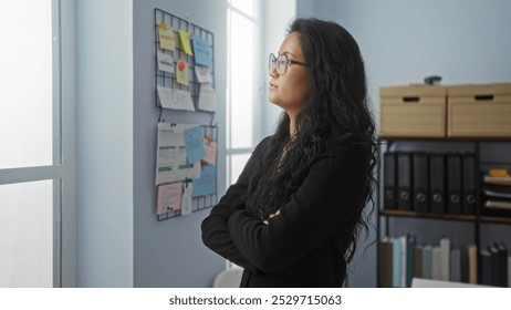 Woman with long black hair and glasses standing with arms crossed in an office, staring thoughtfully at a bulletin board with notes and documents, showcasing a professional and focused demeanor - Powered by Shutterstock