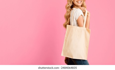 A woman with long blonde hair smiles and holds a white canvas tote bag against a pink background. Her body is turned to the side, and she is only partially visible in the photo, cropped - Powered by Shutterstock