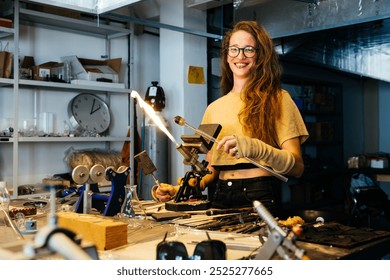 A woman with long brown hair and glasses smiles for the camera while working on a glassblowing project. - Powered by Shutterstock