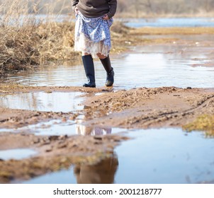 A Woman In A Long Dress And Rubber Boots Crosses Mud And Puddles After A River Flood, A Spring Day