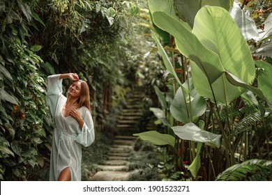 Woman in long elegant silk dress posing in tropical garden, closed eyes, happy and smiling