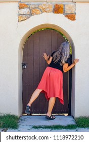 Woman With Long Grey Hair Peeks Around Locked Arched Door In Wall To Garden Beyond