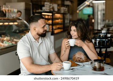 A woman with long hair with brown maltipoo and a bearded man in a cap are talking and eating croissants and drinking tea in cafe. Couple in love with little dog having breakfast in cafe. - Powered by Shutterstock