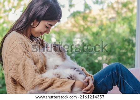 Woman with long hair in brown sweater and jean holding persain cat in her lap and embrassed him with love feelling, girl and cat looking each other with love and care emotion.