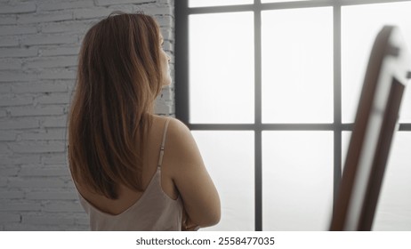 Woman with long hair gazes thoughtfully through a large window from her bedroom, capturing a moment of solitude at home in a serene indoor setting. - Powered by Shutterstock