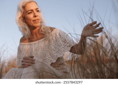 A woman with long hair in a lace dress enjoys a tranquil moment in a natural outdoor environment, surrounded by tall grass, bathed in warm sunlight. - Powered by Shutterstock