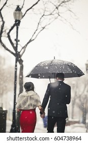 A Woman In A Long Red Evening Dress And A Man In A Suit, Walking Through Snow In The City