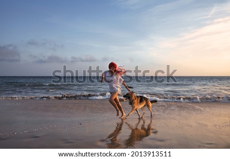 Woman with long red hair running with her German Shepherd dog on a beach at sunset with reflections on the wet sand in a healthy lifestyle concept. Friendship, fun and enjoyment at summer vacation