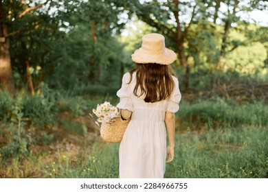 a woman in a long summer dress and a wicker hat on her head walks with her back to the camera along a forest path enjoying a sunny day - Powered by Shutterstock