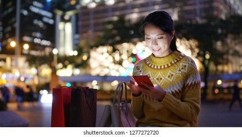 Woman Look At Mobile Phone In City With Shopping Bag At Night