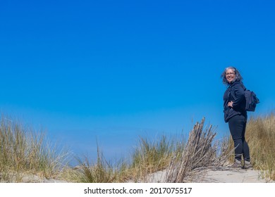 Woman Looking At The Camera With A Blue Background, Standing On A Sandy Hill Of A Dune With Marram Grass, Grayish Black Hair In Black Hiking Clothes With A Backpack. Brouwersdam, Zeeland, Netherlands