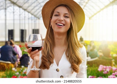 Woman looking at camera and holding glass of wine with people among flowers field drinking and chatting on the background