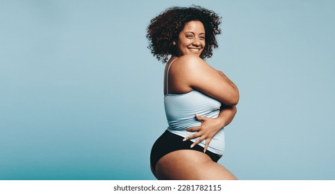 Woman looking at the camera as she proudly embraces her body in its natural form. Woman standing confidently in a fitness studio, dressed in sportswear and radiating self-love and acceptance.