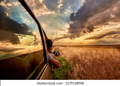 A Woman Looking In The Car At The Sun Set. The Sky Is Full Of Clouds. There Is The Reflection Of The Sky In The Window Of The Car.