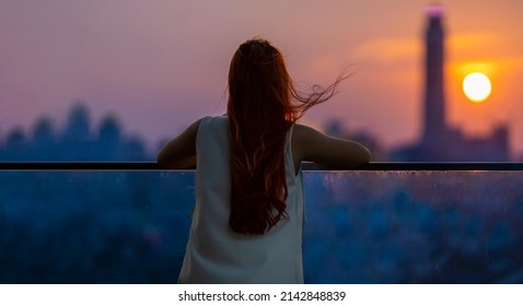 Woman looking and enjoying the sunset view from balcony with the sun setting behind skyscraper in busy urban downtown with loneliness for solitude, loneliness and dreaming of freedom lifestyle concept