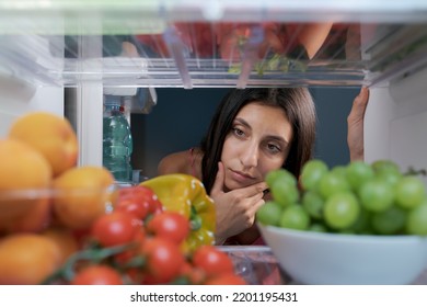 Woman Looking In The Fridge And Thinking With Hand On Chin, Fresh Fruit And Vegetables In The Foreground, POV Shot From Inside Of The Fridge