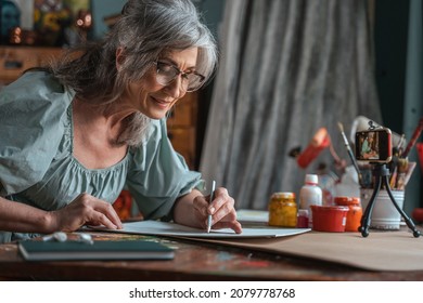 Woman looking at her painting at home while drawing and pencil in front the smartphone