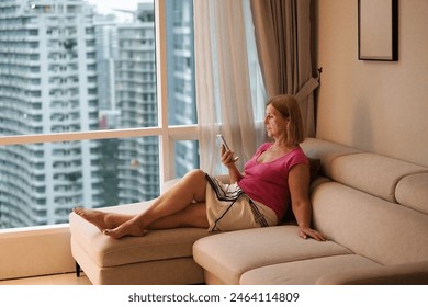 Woman looking at her phone at big window with city view. Female enjoying calm quiet time with social media. People read and relax. Modern apartment in high rise building. - Powered by Shutterstock