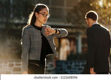 Woman Looking At Her Wrist Watch While Commuting To Office In The Morning. Woman Going To Office Checking Time While Walking On Street With Sun Flare In The Background.