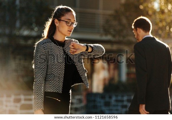 Woman looking at\
her wrist watch while commuting to office in the morning. Woman\
going to office checking time while walking on street with sun\
flare in the background.