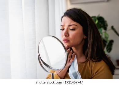 Die Frau sieht sich im Spiegel zu Hause an dem großen Fenster an. Sie macht sich Sorgen um Akne, Maskne