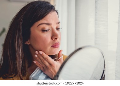 Woman Looking Herself In The Mirror At Home Standing By The Big Window. She Is Concerned About Acne, Maskne