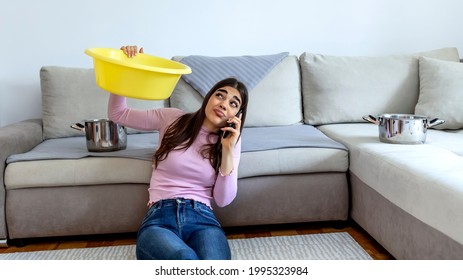 Woman looking up and holding pot under leaking ceiling. Irritated woman sitting on sofa with silver pot. Upset girl calling plumber. Female calling roof repair service while water leaking from ceiling