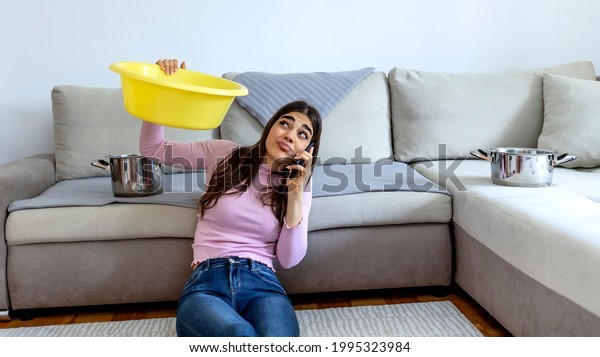 Woman looking up and holding pot under leaking\
ceiling. Irritated woman sitting on sofa with silver pot. Upset\
girl calling plumber. Female calling roof repair service while\
water leaking from\
ceiling