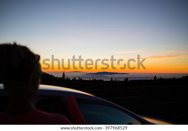 Woman looking at inspirational\
landscape and ocean view. Female car driver or passenger silhouette\
and beautiful sunset sky on Tenerfie Canary Islands,\
Spain.