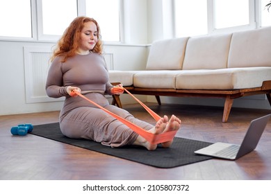 Woman Is Looking Into Computer While Doing An Exercise In Comfortable Living Room, Sitting On Fitness Mat, Overweight Lady In Sprotive Outfit Is Engaged In Sport. Wellbeing, Endurance, Weight Loss