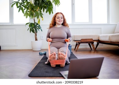 Woman Is Looking Into Laptop While Doing An Exercise In Comfortable Living Room, Sitting On Fitness Mat, Overweight Lady In Sprotive Outfit Is Engaged In Sport. Wellbeing, Endurance, Weight Loss