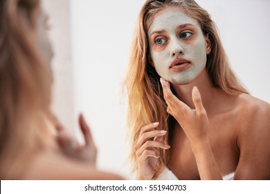 Woman looking in the mirror with mask on her face. Female applying facial cosmetic mask in bathroom.