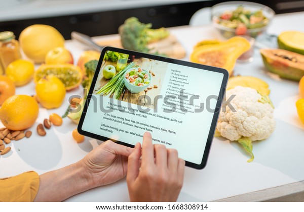 Woman looking on the digital recipe, using\
touchscreen tablet while cooking healthy meal on the kitchen at\
home, close-up view on the\
screen