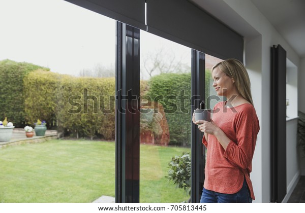 Woman is looking out of her patio door windows\
with a cup of tea in her hands.\
